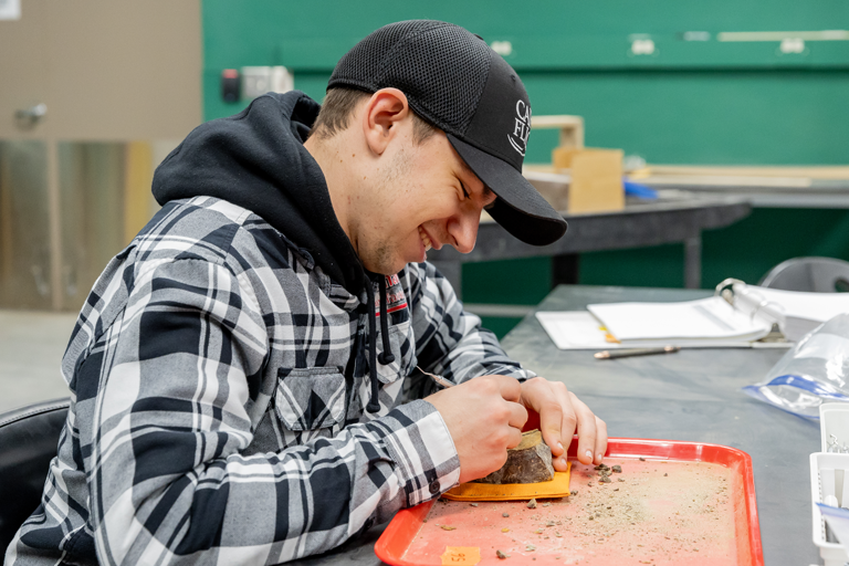 a smiling student works to release a fossil from rock on a tray