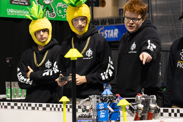 two students with pineapple hats study a robot on the competition field while another boy points