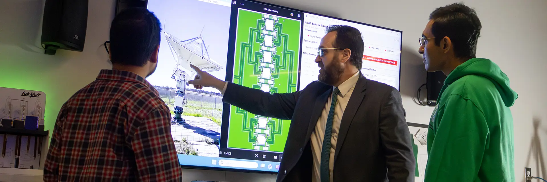 man in suit points to radar image on screen as two students look on