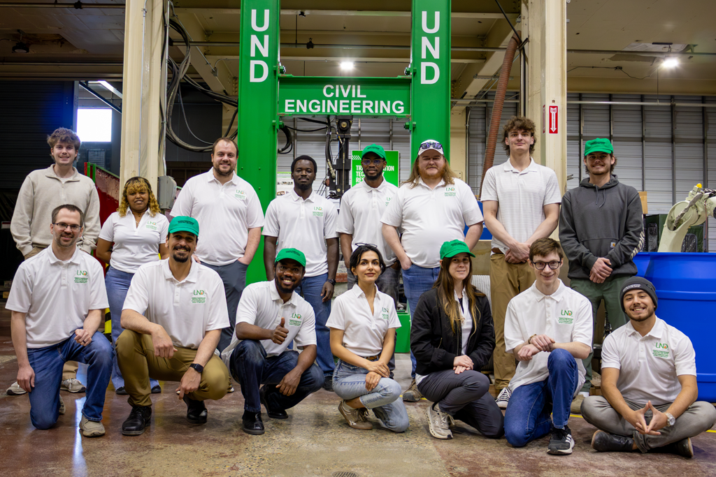a group of people in white shirts, many with green ballcaps, pose in front of a piece of equipment labled "UND Civil Engineering"