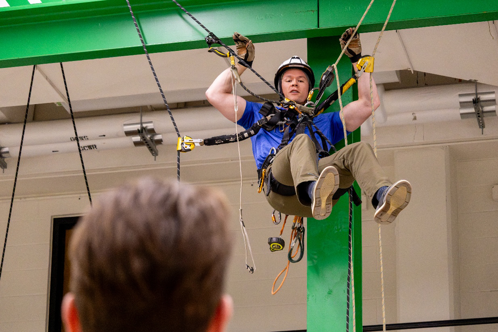 a man in a harness and hard hat hangs from a green beam