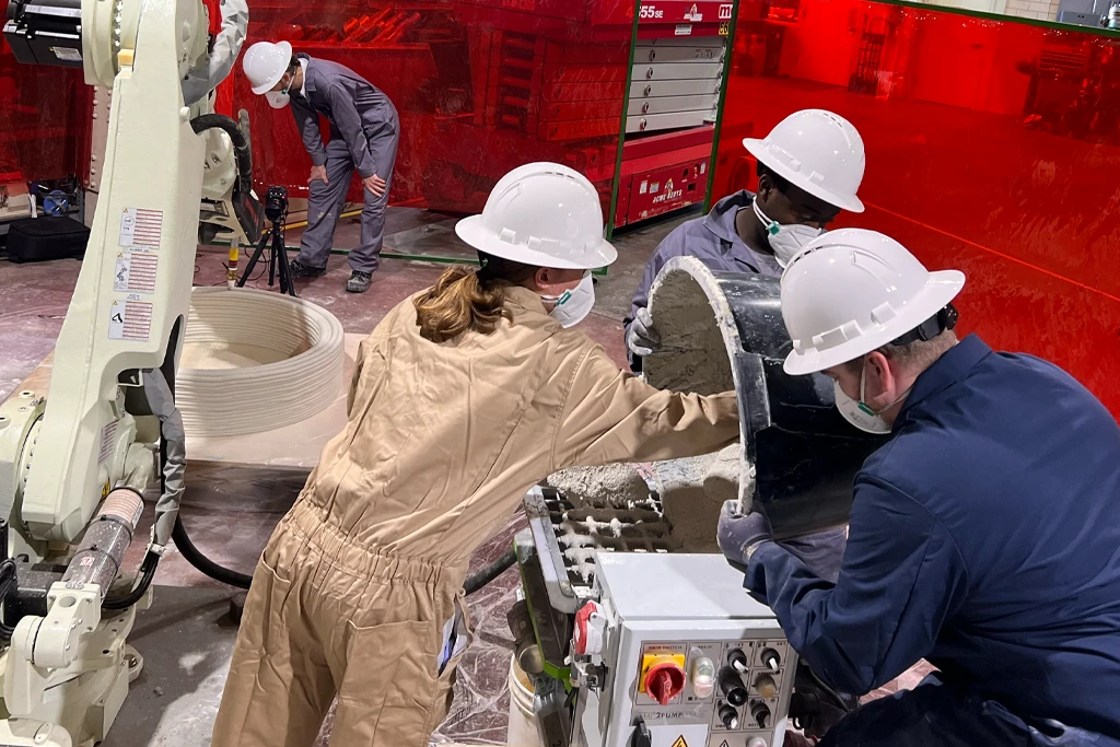 four people work with a 3D concrete printer, a red screen provides protection