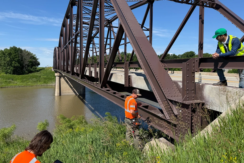 Three people in high-visibility vests inspect a dark brown steel truss bridge over a river on a clear day. One person leans on the bridge railing taking notes, another stands on the embankment beside a riveted girder, and a third works in the foreground grass.