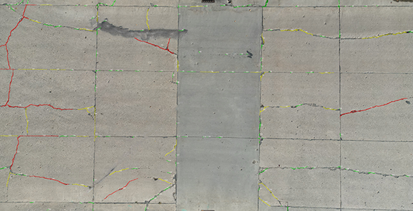 Overhead image of a jointed concrete surface with cracks traced in colored lines. Red lines mark wide cracks, yellow lines mark narrower cracks, and green lines follow the straight construction joints between panels.