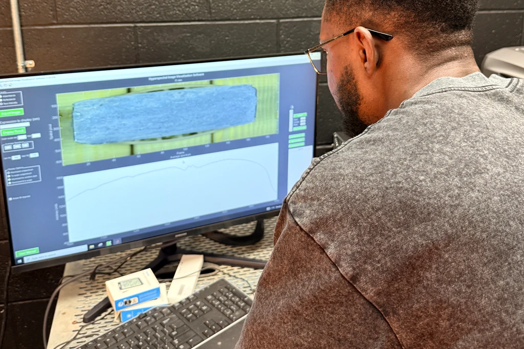 A student seated at a keyboard looks at a widescreen monitor showing hyperspectral acquisition software. The upper pane displays a false-color scan of a long rectangular specimen and the lower pane shows a spectral reflectance plot.