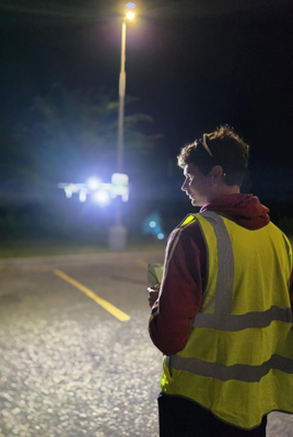 Alt text: A crew member in a yellow high-visibility vest stands on a parking lot at night holding a controller, watching a multirotor drone hover in the distance with bright white navigation lights lit beneath a streetlight.