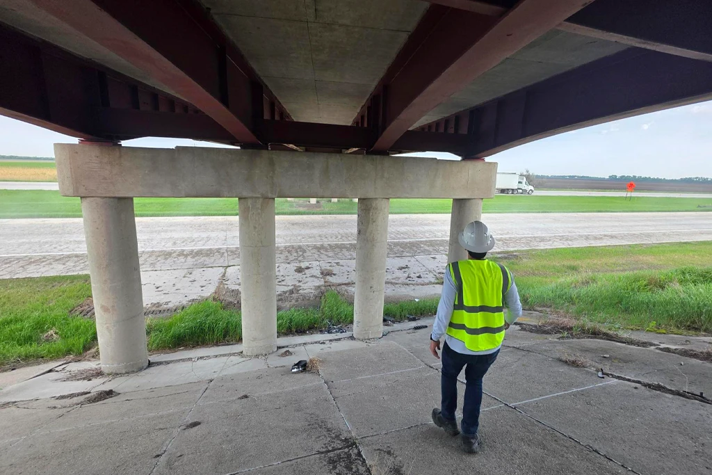 View from beneath a highway bridge showing the concrete deck underside, brown steel girders, and a concrete pier cap on round columns. A researcher in a white hard hat and yellow safety vest walks toward the roadway, where a semi-truck passes in the distance.