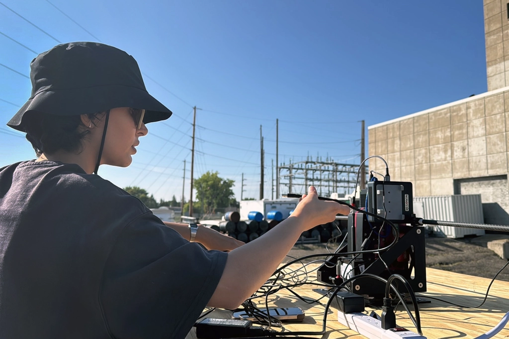 A researcher in a black bucket hat and sunglasses reaches toward a black-framed sensor rig on a plywood table outdoors, plugging in a cable. The rig holds a Resonon hyperspectral camera and a carbon-fiber boom supporting a GNSS antenna puck. An electrical substation, power lines, and a concrete industrial building are in the background under a clear blue sky.