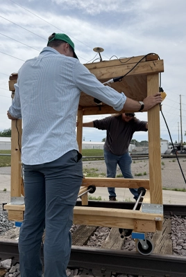 Two people position a wooden-framed cart on a railroad track. One, in a striped shirt and green cap, secures a component to an upright post while the other, in a brown shirt, steadies the far side of the frame. A GNSS antenna and cabling are mounted on top, and rubber caster wheels ride on the rails above the ballast and ties.